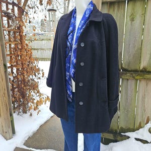 May include: A black wool coat with a classic design, featuring a button-up front and a collared neckline. A blue scarf with a white pattern is draped over a white turtleneck. The coat is paired with blue jeans.