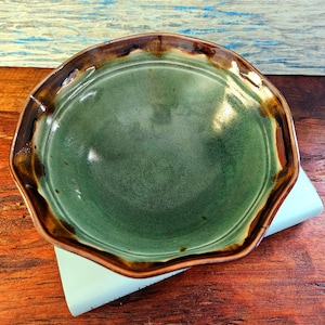 May include: A handmade ceramic plate with a unique, irregular shape. The plate features a deep green interior and a contrasting brown rim. The plate is resting on a light blue surface, showcasing its handcrafted nature.