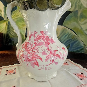 Könnte beinhalten: Eine weiße Keramikkrug mit einem rosa Blumenmuster. Der Krug hat einen gebogenen Griff und einen Ausguss. Er steht auf einem weißen Leinenläufer mit gestickten Details. Grünes Laub befindet sich im Inneren des Kruges.
