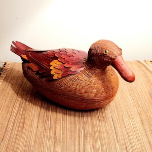 May include: A handcrafted duck figurine made of woven brown material with red and orange accents on the wings. The duck has a long, red beak and golden eyes, and is resting on a woven mat.