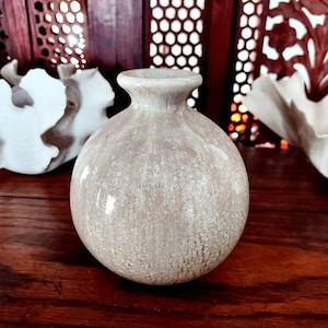 May include: A small, round, white ceramic vase with a textured surface. The vase has a slightly off-white color and a subtle speckled pattern.