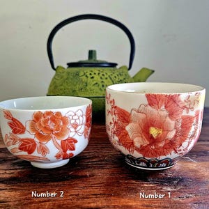 May include: Two ceramic tea cups with floral designs in shades of orange and red. The cups are white with a rounded shape. A green cast iron teapot is in the background. The cups are labeled "Number 1" and "Number 2".