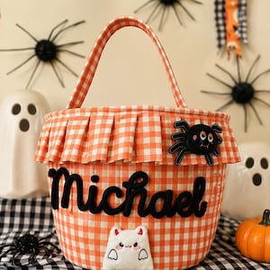May include: An orange and white gingham fabric bucket with a handle, featuring the name Michael in black felt lettering. The bucket has a pleated rim, a black spider applique, and a white ghost applique with small red horns on the front.