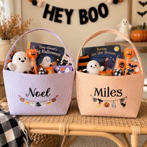 May include: Two fabric Halloween baskets with handles, one lavender and one orange, both featuring fine vertical stripes. Each basket is embroidered with a name, 'Noel' or 'Miles', and a festive garland graphic. They contain plush ghost toys, books, and candy.
