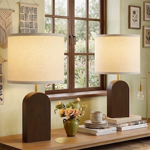 May include: Two matching table lamps with dark brown wooden bases and beige fabric shades. The lamps have a gold-colored metal stem. The lamps are on a wooden table with books, a teacup, and a vase of flowers. The background includes a window and wall art.