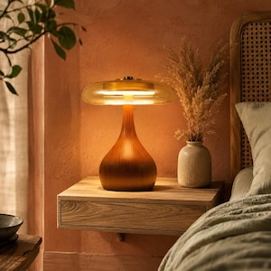 May include: A table lamp with a smooth, teardrop-shaped wooden base and a wide, translucent amber glass shade. The lamp emits a warm, soft glow from an internal light source, illuminating the surrounding area with a gentle, diffused light.