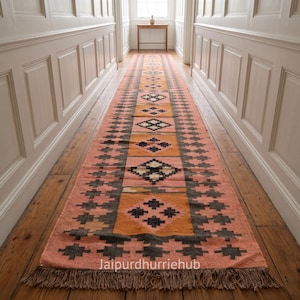 May include: A long runner rug featuring a repeating geometric diamond pattern in shades of peach, orange, and dark charcoal grey. The rug has a fringed edge at the bottom and displays the text Jaipurdhurriehub printed near the lower border.