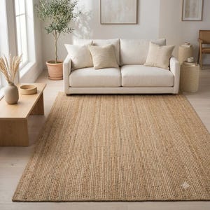 Puede incluir: Una alfombra rectangular hecha de fibras de yute tejidas naturales en un color tostado claro. La alfombra presenta una textura consistente y de tejido apretado en toda su superficie, proporcionando una base neutra que cubre el suelo frente a un sofá.