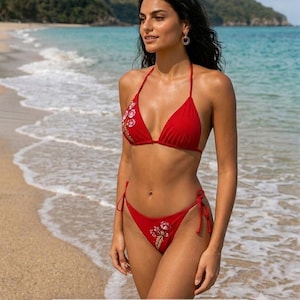 May include: A vibrant red two-piece bikini featuring a triangle top and matching side-tie bottoms. Both pieces are adorned with delicate floral embroidery in shades of pink, white, and green, adding a detailed accent to the solid red fabric.