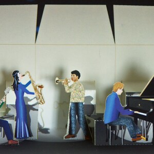 May include: A paper cutout diorama of a jazz band performing on a stage. The band includes a guitarist, a saxophonist, a trumpet player, and a pianist playing a black grand piano. The background is a light gray.