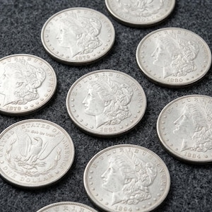 Könnte beinhalten: Eine Sammlung silberfarbener Morgan-Dollar-Münzen mit dem Profil der Lady Liberty auf der Vorderseite, datiert auf 1879, 1880, 1881, 1884 und 1894, sowie eine Münze, die die Rückseite mit einem Adler und dem Text United States of America zeigt.