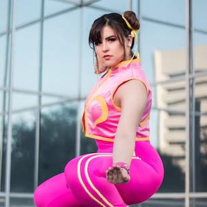 Puede incluir: Cosplay de Chun-Li con top corto rosa, leggings y zapatillas deportivas. El atuendo presenta detalles amarillos. La persona está en posición de combate, con expresión seria. El fondo es un edificio moderno.