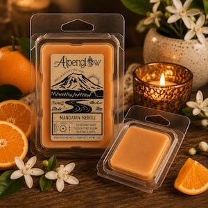 May include: Two clear plastic clamshell packages containing orange wax melts. The larger package features a label with the text Alpenglow, Mandarin Neroli, and a mountain illustration. The wax is a solid orange color, and the net weight is 2.4 oz.