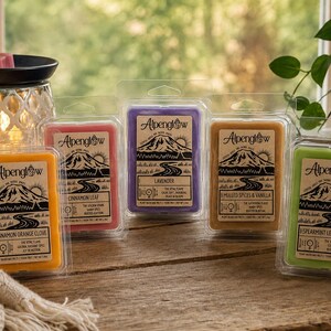 May include: Five rectangular wax melt packages labeled Alpenglow with mountain illustrations, featuring scents Cinnamon Orange Clove, Cinnamon Leaf, Lavender, Milled Spices & Vanilla, and Spearmint Lemongrass, arranged near a lit wax warmer.
