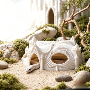 May include: A white, 3D-printed reptile hide shaped like a miniature gothic castle with pointed turrets, arched doorways, and intricate fan-vaulting details. A pale, smooth-scaled snake is partially emerging from the main arched entrance.