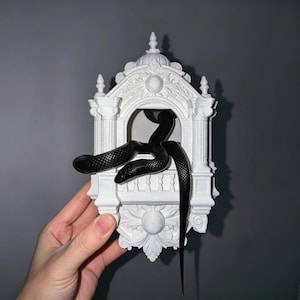 May include: A white 3D printed architectural structure featuring a central arched opening with a balcony, adorned with ornate columns and decorative carvings, with a long, smooth, black snake coiled through the opening.