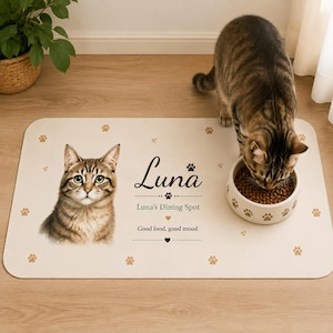 Puede incluir: Un tapete rectangular de color blanco hueso para mascotas con una ilustración de un gato atigrado marrón, huellas de patas y el texto "Luna", "Luna's Dining Spot" y "Good food, good mood". Un cuenco con pienso se encuentra sobre la superficie.