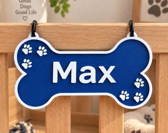 May include: A bone-shaped sign in deep blue with a white border and the name Max in white lettering. The sign features small white paw prints on the left and right sides and includes two black metal hooks at the top for hanging on a wooden surface.