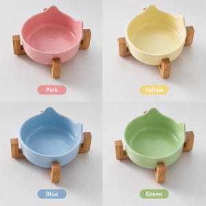 May include: Four ceramic bowls with cat-ear shaped rims, each resting on a light brown wooden stand. The bowls are available in pastel pink, yellow, light blue, and light green. Each bowl is labeled with its corresponding color name below it.