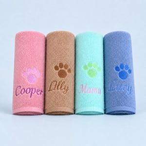May include: Four rolled towels in pink, tan, light blue, and periwinkle blue. Each towel features an embroidered paw print and a name: Cooper, Lilly, Mamu, or Lucy. The embroidery colors match the paw prints and coordinate with the towel colors.