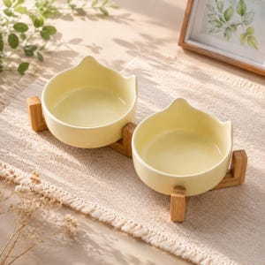May include: Two pale yellow ceramic bowls with cat-ear shaped rims, each resting on a separate light-colored wooden stand. The bowls have a smooth, glossy finish and are designed for feeding, featuring a simple, functional structure.