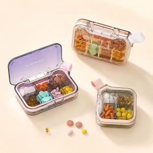 Può includere: Tre portapillole in plastica trasparente con più scomparti, ciascuno riempito con pillole e capsule di vari colori. I portapillole hanno dettagli rosa e trasparenti, con una maniglia laterale. Il testo "STORAGE BOX" è visibile.