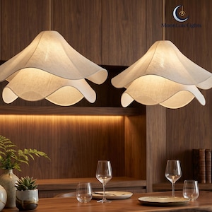 May include: Two pendant lights with a layered, petal-like design hang above a wooden surface. The lampshades are a light beige color, and the room features wooden cabinetry and a table set with glassware and plates. The logo "MoonLureLights" is visible.