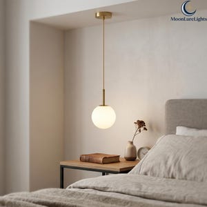 May include: A modern pendant light with a gold finish, featuring a spherical, frosted glass shade. The light hangs from a thin gold rod, illuminating a bedroom scene with a neutral color palette. The text "MoonLureLights" is visible in the upper right corner.