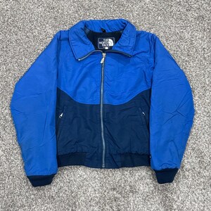 May include: A bright blue and dark navy blue zip-up jacket featuring a high collar and ribbed cuffs. The jacket has a central silver zipper and a black interior label with white text reading The North Face. Two zippered pockets are on the front.