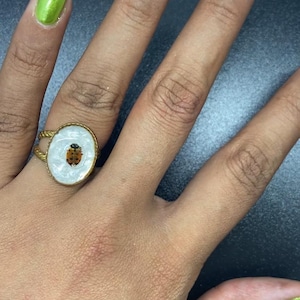 May include: A gold-toned ring featuring an oval-shaped setting with a white, pearlescent inlay and a small ladybug graphic at the center. The ring band has a twisted metal texture, and the piece is worn on a finger with lime green and white nail art.