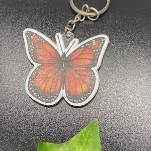 May include: A monarch butterfly keychain featuring a flat, die-cut acrylic charm with orange and black wing patterns, white borders, and a silver-toned metal split ring attached by a small chain link, showcasing detailed insect wing veins.