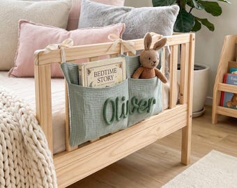 Peut inclure: Une pochette de rangement de chevet en tissu vert sauge suspendue à un cadre de lit en bois clair. La pochette porte le nom Oliver en lettres vertes en relief, contenant un livre intitulé Bedtime Story et un petit lapin en tricot marron.
