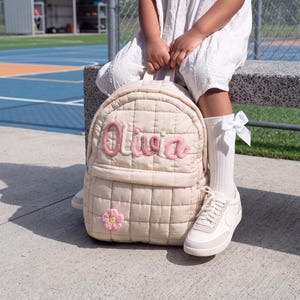 May include: A cream-colored quilted backpack featuring a grid pattern and the name Olivia embroidered in pink script across the front pocket. A small pink and yellow flower applique is attached to the lower left corner of the backpack.