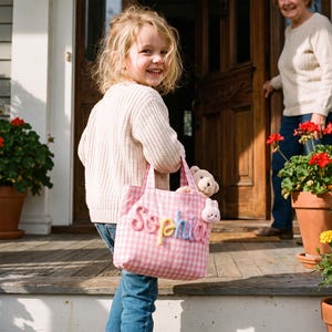 May include: A pink and white gingham tote bag featuring the name Sophia in raised, multi-colored fabric lettering. The bag has two fabric handles and contains a small, light brown plush teddy bear tucked inside the main compartment.