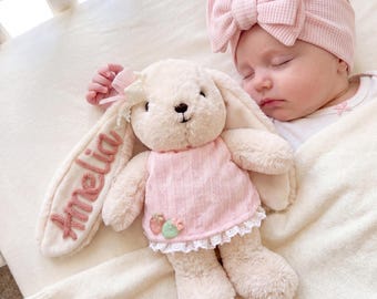 May include: A plush cream-colored bunny toy wearing a pink textured dress with white lace trim and two small fruit charms. The bunny has the name Amelia embroidered in pink thread on one long ear. A baby wears a matching pink waffle-knit headband.