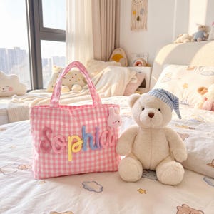 May include: A pink and white gingham tote bag featuring the name Sophia in multi-colored yarn lettering on the front, accompanied by a small pink plush bunny charm attached to the handle, sitting next to a cream-colored teddy bear wearing a blue striped hat.
