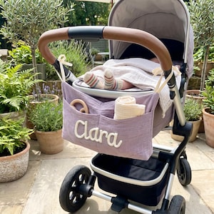 May include: A mauve fabric pram organiser attached to a grey baby carriage with a brown handle. The organiser features the name Clara in white script lettering, a wooden ring, and a folded white cloth. A baby is visible inside the carriage.