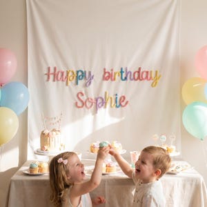 May include: A white fabric backdrop featuring the text Happy birthday Sophie in colorful yarn lettering. The scene includes a birthday cake, cupcakes, and pastel balloons in pink, blue, and yellow, arranged on a table covered with a neutral cloth.
