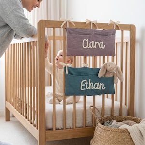 Peut inclure: Deux poches de rangement rectangulaires en tissu suspendues &agrave; un lit &agrave; barreaux en bois. La poche sup&eacute;rieure est violette avec le pr&eacute;nom Clara brod&eacute; en blanc. La poche inf&eacute;rieure est bleu sarcelle avec le pr&eacute;nom Ethan et un lapin en peluche.