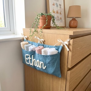 Peut inclure: Un organisateur de rangement suspendu en tissu bleu &agrave; trois poches, avec le pr&eacute;nom Ethan brod&eacute; en &eacute;criture blanche. L'organisateur est attach&eacute; &agrave; une commode en bois avec des rubans cr&egrave;me et contient six langes en mousseline roul&eacute;s.