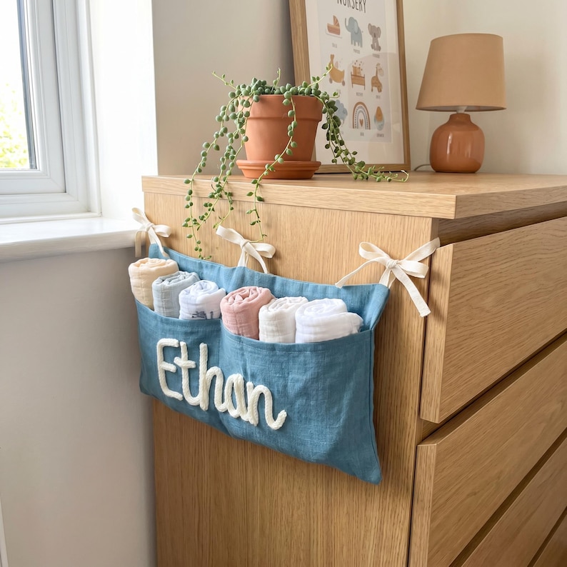 Peut inclure: Un organisateur de rangement suspendu en tissu bleu &agrave; trois poches, avec le pr&eacute;nom Ethan brod&eacute; en &eacute;criture blanche. L'organisateur est attach&eacute; &agrave; une commode en bois avec des rubans cr&egrave;me et contient six langes en mousseline roul&eacute;s.