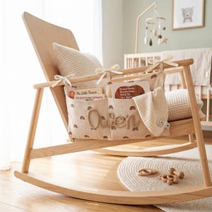 Peut inclure: Un organisateur de rangement en tissu beige clair avec un motif r&eacute;p&eacute;titif d'arcs-en-ciel marron et le nom Owen en lettres cursives en relief, attach&eacute; &agrave; l'accoudoir d'un fauteuil &agrave; bascule en bois clair avec des rubans cr&egrave;me, contenant deux livres.