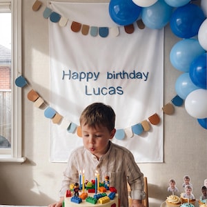 May include: A white fabric banner with the blue text Happy birthday Lucas, decorated with a garland of blue, tan, and white semi-circular felt pieces. A cluster of blue and white balloons is attached to the right side of the banner.
