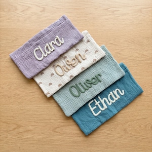 Peut inclure: Quatre pochettes rectangulaires en tissu mousseline froiss&eacute;. Les pochettes sont de couleur violette, cr&egrave;me avec un motif arc-en-ciel marron, vert sauge et bleu sarcelle. Chaque pochette porte un nom brod&eacute; en &eacute;criture cursive : Clara, Owen, Oliver et Ethan.