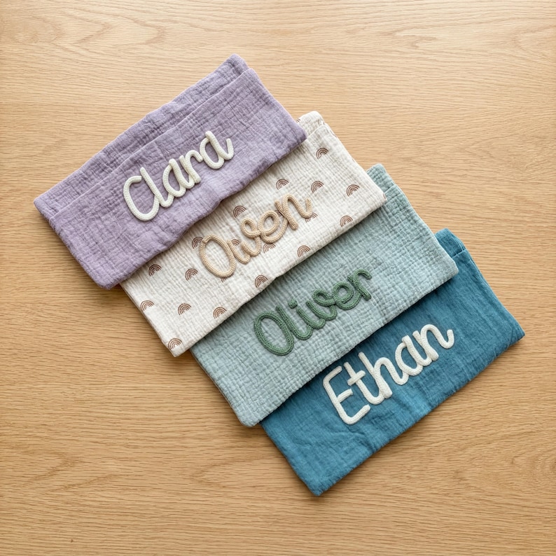 Peut inclure: Quatre pochettes rectangulaires en tissu mousseline froiss&eacute;. Les pochettes sont de couleur violette, cr&egrave;me avec un motif arc-en-ciel marron, vert sauge et bleu sarcelle. Chaque pochette porte un nom brod&eacute; en &eacute;criture cursive : Clara, Owen, Oliver et Ethan.