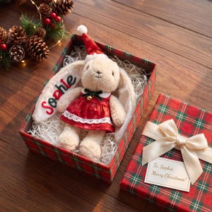 May include: A plush cream-colored rabbit toy wearing a red dress with white snowflake patterns and a matching Santa hat. The rabbit's ear is embroidered with the name Sophie in red and green. It sits in a red and green plaid gift box.