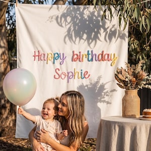 May include: A white fabric backdrop featuring the text Happy birthday Sophie in colorful, raised, rounded lettering. The banner is displayed outdoors, accompanied by a pastel iridescent balloon, a woven vase with dried flowers, and a small cake.