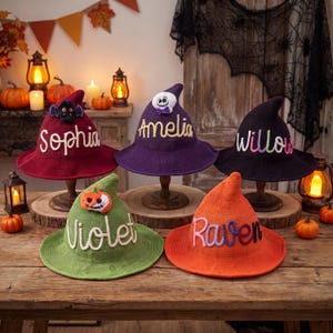 May include: Five knitted witch hats in maroon, purple, black, green, and orange. Each hat features a unique embroidered name: Sophia, Amelia, Willow, Violet, and Raven. Decorative felt accents include a bat, a skull, and a pumpkin.