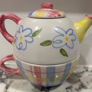 Op de afbeelding: Een keramische thee-voor-één set bestaande uit een theepot gestapeld op een theekopje. De theepot heeft een witte basis met handgeschilderde lichtblauwe bloemen en groene blaadjes. Het deksel en de onderkant van de kop hebben een kleurrijk ruitpatroon.