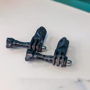 Puede incluir: Dos adaptadores de montaje para cámara de plástico negro con juntas de tres puntas y tornillos de mariposa metálicos integrados. Estos conectores en forma de L presentan un acabado texturizado en las pestañas de montaje y un pomo.